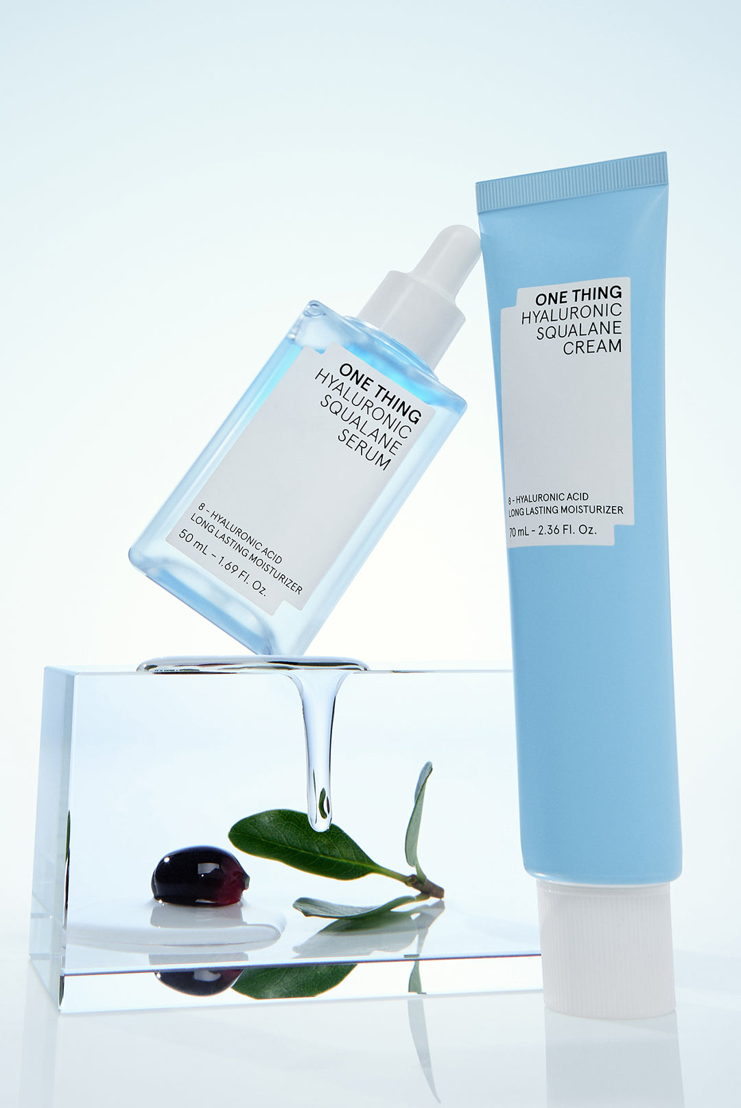 Two skincare products, a serum and a cream, with one standing upright and the other lying down, beside a glass block and a leaf, on a white background.