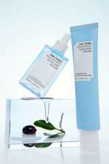 Two skincare products, a serum and a cream, with one standing upright and the other lying down, beside a glass block and a leaf, on a white background.